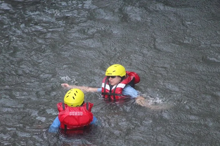 Two people wearing life jackets and helmets swim in a murky river.