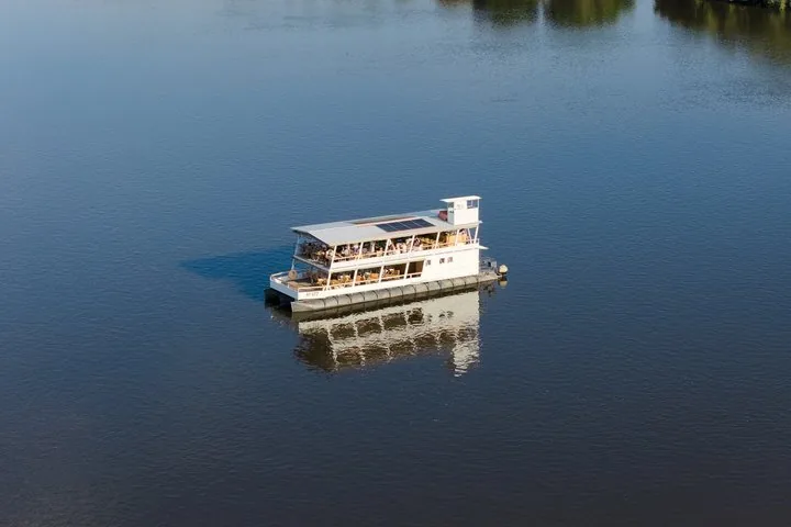 Ein zweistöckiges weißes Boot gleitet bei klarem Himmel über einen ruhigen blauen Fluss.